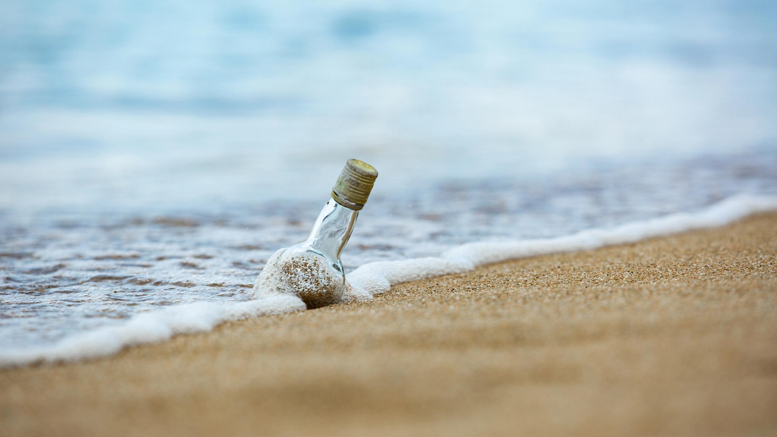 Glasflasche mit Korken im Sand am Strand, umspült von Schaum der Brandung